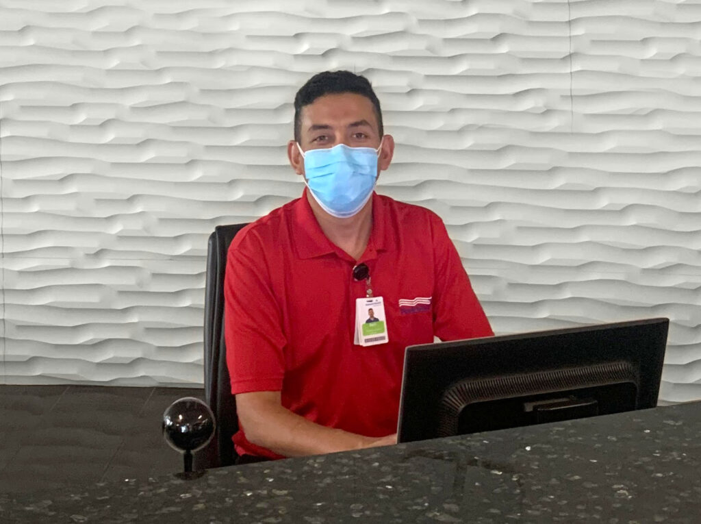 man sitting behind a desk with the top of the computer screen in view. He is wearing a red ParkMed Hospitality mnaager uniform polo and a blue mask over his mouth and nose.